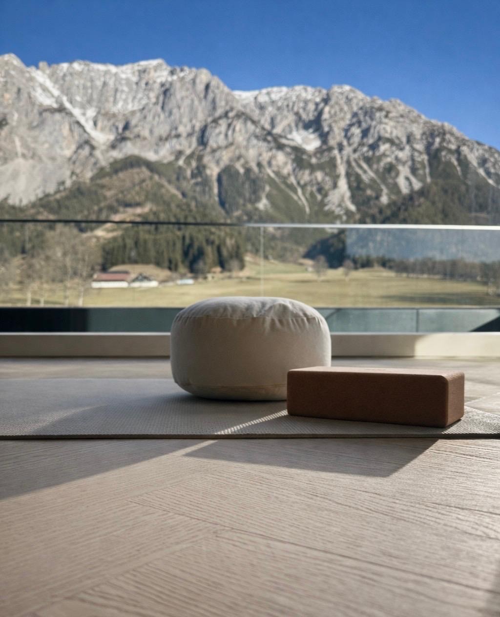 Yogamatte mit Meditationskissen vor Panoramafenster mit Blick auf alpine Berglandschaft